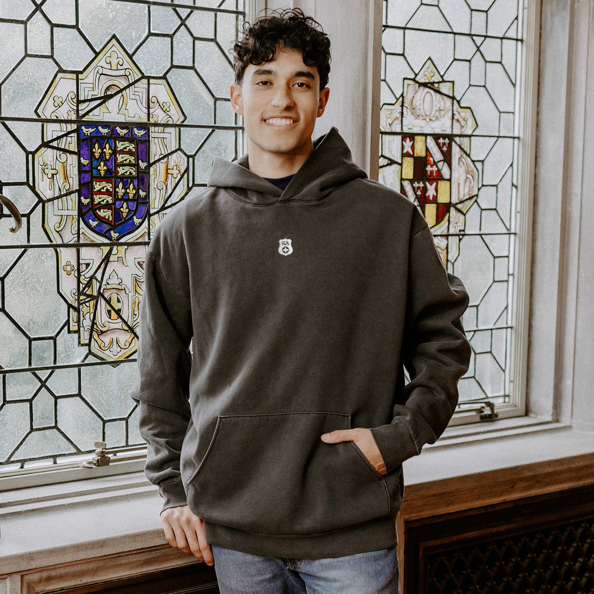 College-aged man wearing the Kappa Alpha embroidered black hoodie, standing in front of stained glass windows with fraternity-style crest designs.
