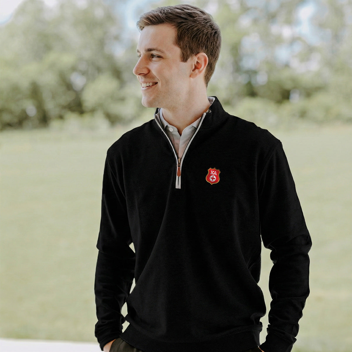 College-aged man smiles and looks off to the side while wearing a black quarter-zip pullover embroidered with a fraternity badge. He stands outdoors in a bright, grassy setting.