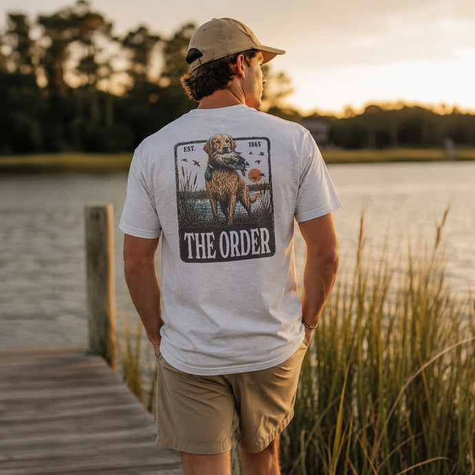 Young man wearing a white graphic T-shirt while standing on a wooden dock overlooking a lake at sunset.