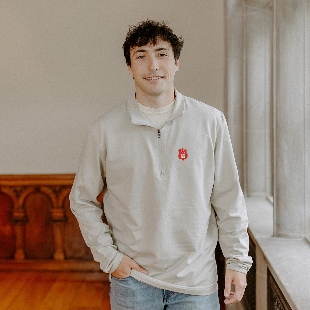 Male model wearing the smoke-colored Kappa Alpha Embroidered Luxe Quarter Zip with Symbol embroidery, standing by a window in a formal setting.