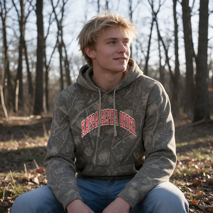 College-aged male wearing the fraternity Deadwood Camo Hoodie with red and white Kappa Alpha Order lettering, seated outdoors in a wooded setting.