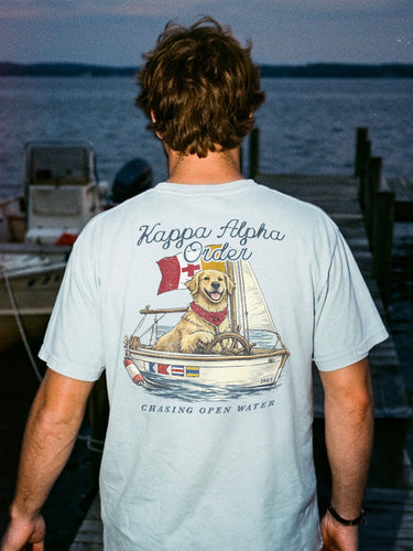 Young man standing on a dock at sunset wearing a light blue t-shirt featuring a golden retriever steering a sailboat with a flag and the phrase “Chasing Open Water” on the back.