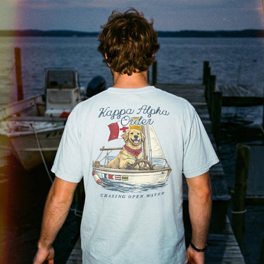 Young man standing on a dock at sunset wearing a light blue t-shirt featuring a golden retriever steering a sailboat with a flag and the phrase “Chasing Open Water” on the back.