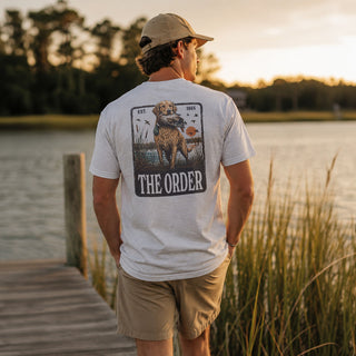 Young man wearing a white graphic T-shirt while standing on a wooden dock overlooking a lake at sunset.
