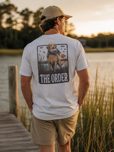 Young man wearing a white graphic T-shirt while standing on a wooden dock overlooking a lake at sunset.