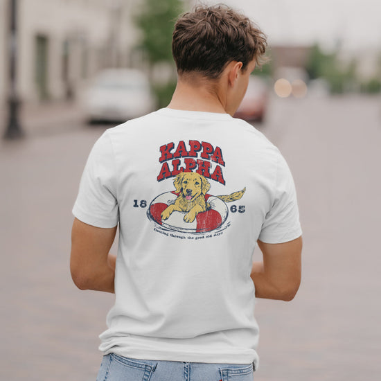 Kappa Alpha Order  fraternity member in a limited edition  white short sleeve tee  featuring a golden retriever puppy playing in an pool floatie  with the fraternity name written above and the founding year.