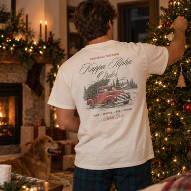 College-aged man wearing a limited edition Tree Farm Holiday Tee in a cozy Christmas setting with a decorated tree and fireplace.