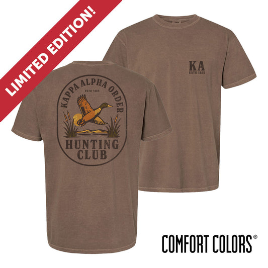 Brown garment-dyed short-sleeve tee featuring a vintage-inspired graphic: a weathered oval frames a mallard in mid-flight with burnt orange and mossy green-tipped wings above rippling water and cattails; distressed Kappa Alpha arcs overhead and Kappa Alpha letters bold and rugged on the front.